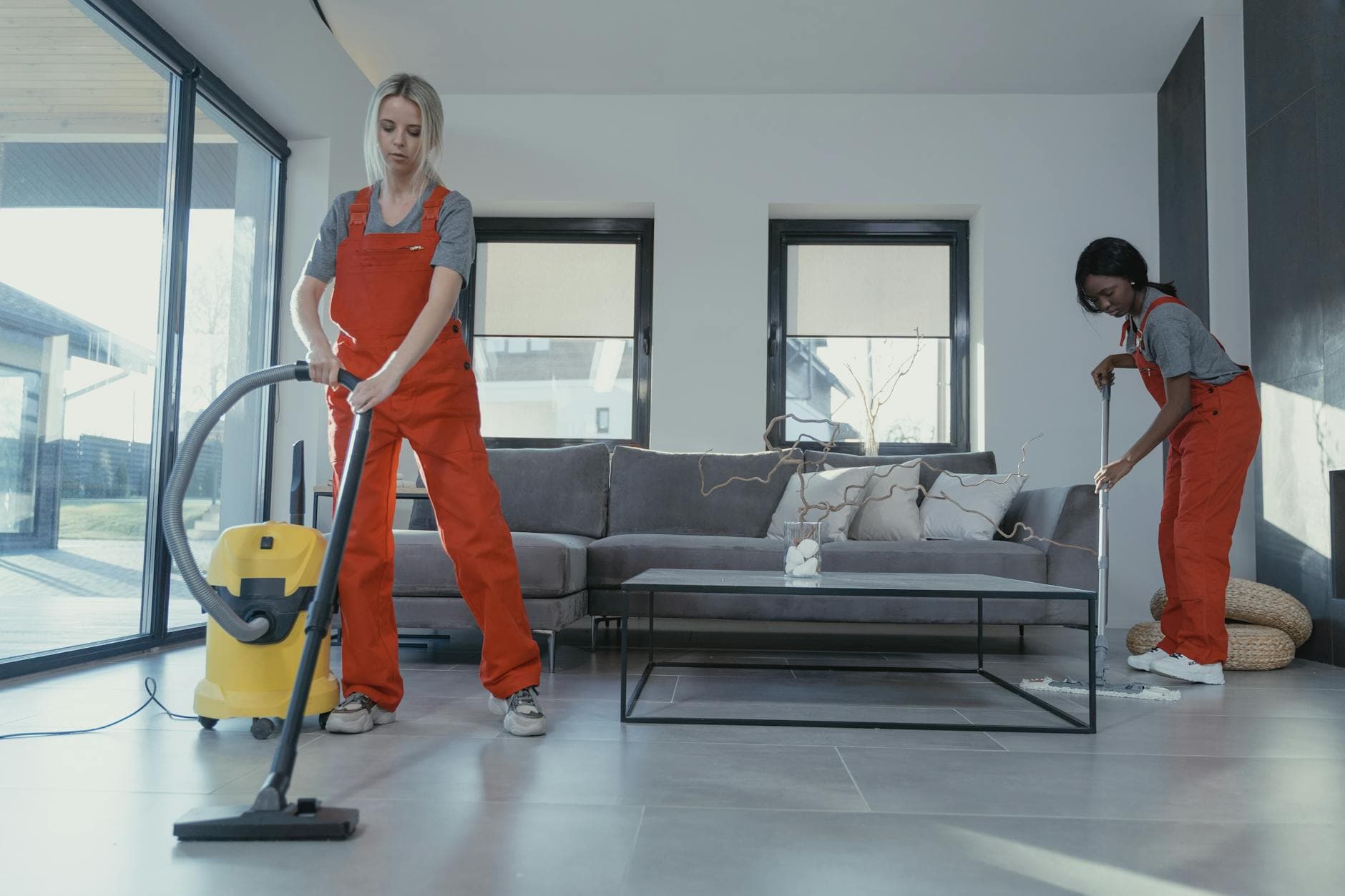 Two NYC Maid cleaners in red overalls vacuuming and sweeping a modern NYC living room — hiring in Westchester Square, Bronx